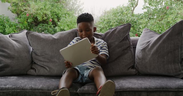 Young African American boy sitting on a modern gray sofa, holding and using a tablet. Ideal for illustrating concepts related to technology use, child-friendly gadgets, childhood leisure activities, and modern lifestyle. Perfect for use in educational content, technology reviews, family-related blogs, and advertisements promoting digital products for children.