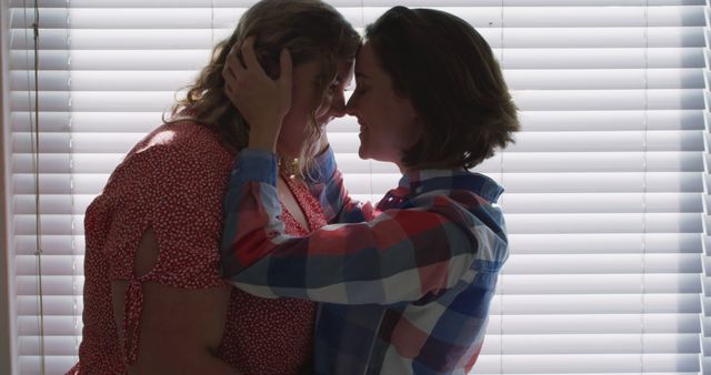 Couple is embracing tenderly while facing each other in front of a window with blinds, with natural light coming from behind creating a backlit effect. This image can be used for themes of romance, relationship, and affection. It is suitable for websites, blogs, and advertisements focusing on intimate moments or love stories.