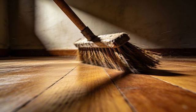 Sunlit broom sweeping hardwood floor, dust motes flying across polished parquet planks