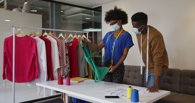 Fashion designers, a man and a woman, are discussing clothes in a workshop. The woman is holding a green dress while the man looks on. They are wearing casual clothes and masks, and sewing supplies like threads and measurements are on the table. The image depicts collaboration, creativity, and health precautions in a modern workspace. Ideal for use in content related to fashion design, teamwork, pandemic precautions, or creative industries.