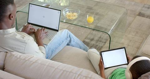 Parent and child using laptop and tablet on sofa learning, streaming, relaxing at home
