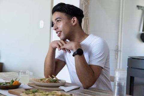 African american man leaning and smiling while eating noodles with chopsticks at minimalist dining t