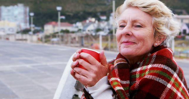Senior woman relaxing outdoors, holding a hot drink and covered in a plaid blanket. Perfect for topics related to senior lifestyle, outdoor activities, relaxation, winter warmth, and health benefits of hot beverages. Ideal for use in blogs, articles, advertisements, and social media promoting comfort and well-being for older adults.