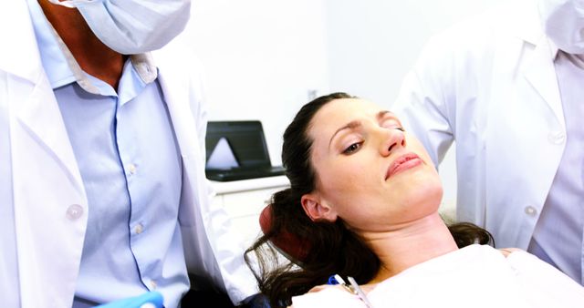 A Caucasian woman is seen reclining in a dental chair, attended to by a dentist and a dental assistant, both wearing surgical masks, with copy space. Their professional attire and the clinical setting suggest a routine dental examination or procedure.