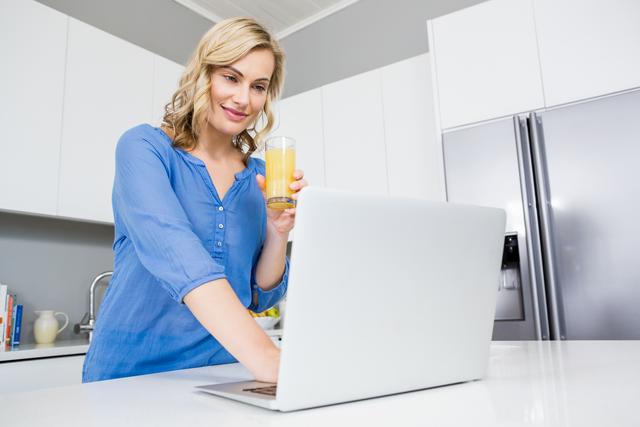 Beautiful woman holding a glass of juice and using laptop in kitchen at home