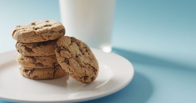 Stack of chocolate chip cookies on white plate, accompanied by glass of milk. Blue background creates clean, appealing look. Perfect for use in food blogs, recipe websites, bakery promotions, and advertisements for dairy or bakery products.