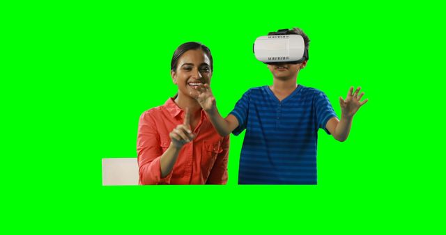A young Asian woman is smiling and gesturing towards a boy wearing a VR headset, both against a green screen background, with copy space. Her enthusiasm suggests she's guiding or teaching the child as he explores a virtual reality environment.