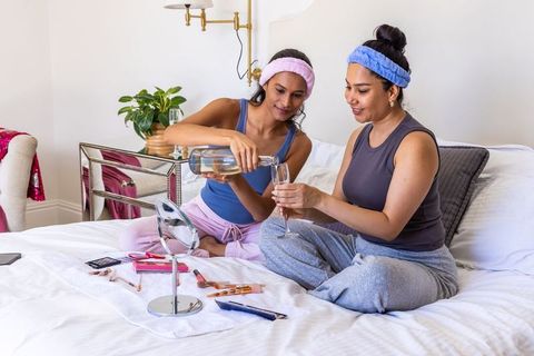 Diverse women enjoying pajama pamper night pouring wine and sharing makeup on bed