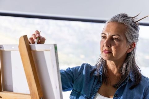 Senior woman painting on canvas in sunlit studio, focused artist at wooden easel