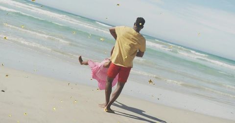 Parent swinging child on sandy beach with gold confetti, playful summer family moment