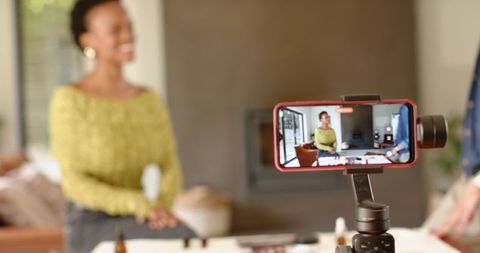 Black woman hosting live beauty demo on smartphone gimbal in cozy living room showcasing skincare