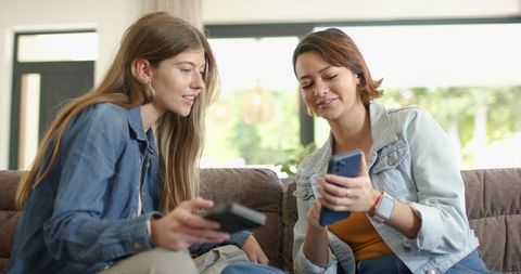 Two women sharing smartphone and holding tv remote on couch in bright living room