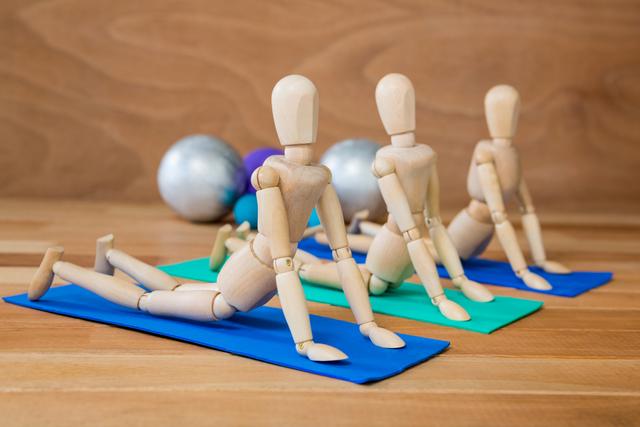 An image of three wooden mannequins on yoga mats performing a stretching exercise. Balls in the background suggest a gym or fitness setting. Ideal for illustrating concepts of fitness, flexibility, and wellness, this image can be used in health and fitness blogs, exercise guides, and wellness promotional materials.
