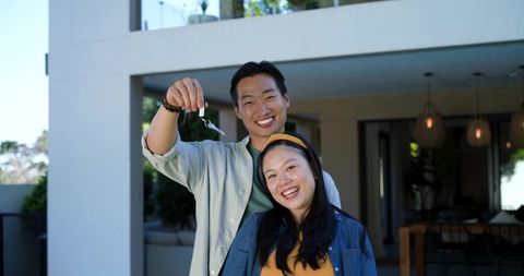 Smiling couple celebrating homeownership, holding keys at modern house entrance