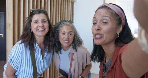 Three friends leaning close, smiling and taking selfie with smartphone in bright modern lobby