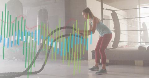 Young woman swinging battle ropes in boxing ring while fitness analytics overlay tracks performance