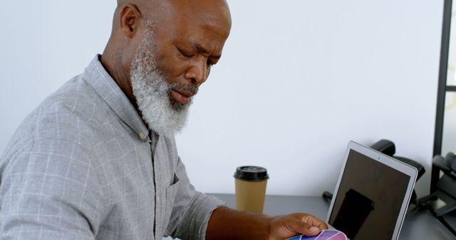 Middle-aged man with beard examining color swatches in modern office. He is sitting at a desk with a laptop and a take-out coffee cup, focusing intently. Ideal for topics related to design, productivity, office work, and creativity.