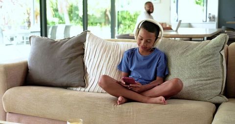 African american father smiling while son listens to music on sofa with headphones