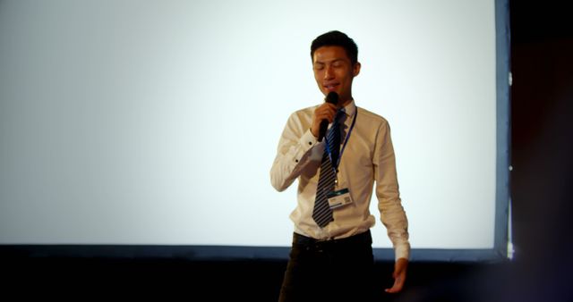 Young male professional speaking into microphone while delivering business presentation on stage. Background shows blank projector screen. Suitable for topics related to business conferences, corporate seminars, public speaking or career advancement tutorials.
