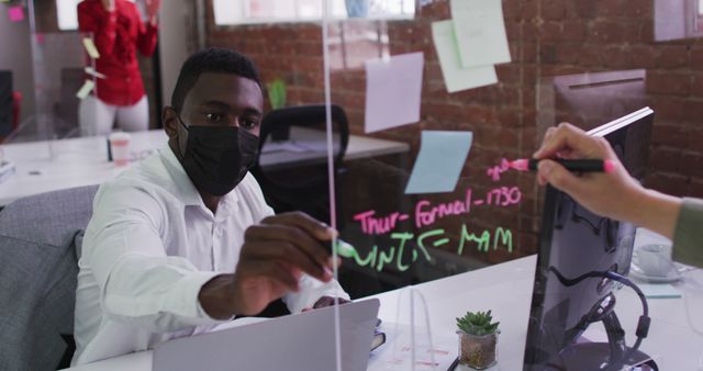 Office worker wearing a mask writing on a glass board with pink and white markers. In the background, colleague in red formal wear. Ideal for illustrating modern workplace, collaboration, pandemic safety measures, and office planning scenes.