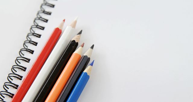 Colored pencils lie next to a spiral notebook on a white background, with copy space. Ideal for educational themes, the image captures the simplicity of art supplies ready for creative work.