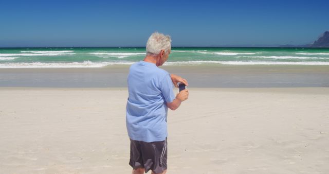 Senior man having vacation by taking photos of ocean waves on a sunny beach. Useful for content focusing on senior activities, travel and tourism, beach relaxation, summer holiday, and outdoor leisure pursuits.