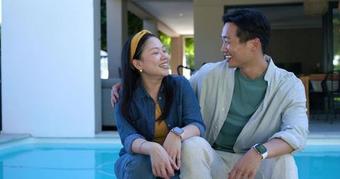 Couple laughing and chatting on pool edge wearing smartwatches, casual outdoor relaxation