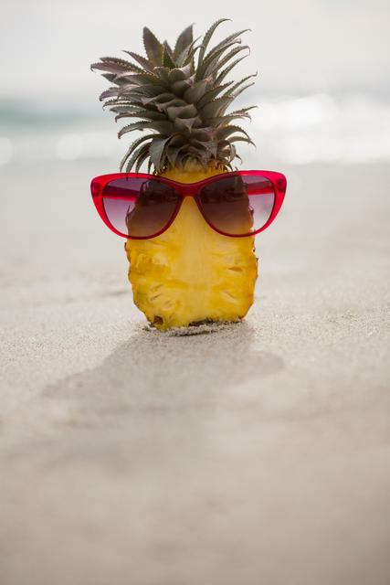 This image shows a halved pineapple wearing red sunglasses on a sandy beach, evoking a fun and playful summer vibe. Ideal for use in travel brochures, summer vacation promotions, tropical-themed party invitations, and social media posts celebrating summer and beach activities.