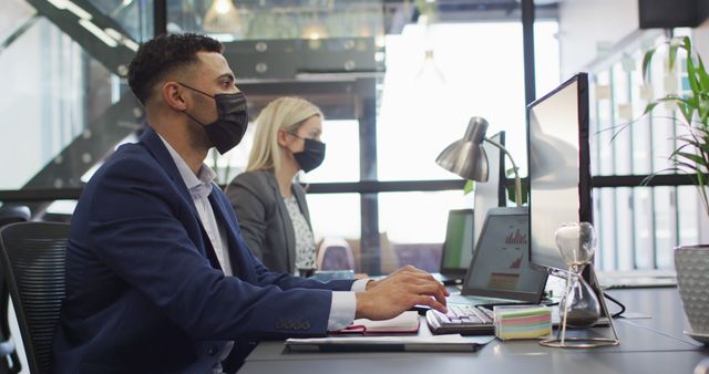 Biracial businessman wearing face mask and using computer with colleague working in background - Download Free Stock Photos Pikwizard.com