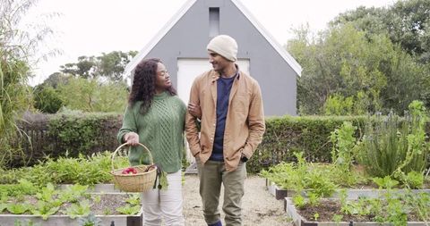 Diverse couple walking arm in arm through cottage garden carrying basket of fresh vegetables