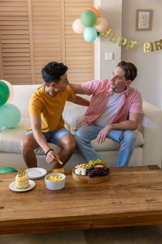 Friends celebrating birthday at home sharing cake and charcuterie on cozy living room sofa