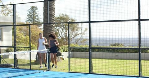 Two women walking onto seaside padel court carrying sports bags on sunny outdoor day