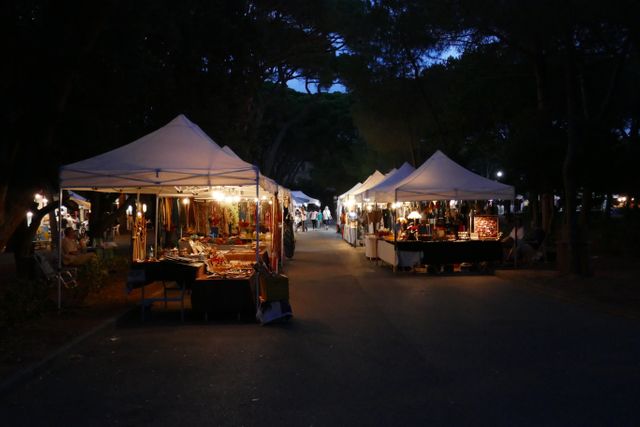 Scene depicts a lively night market with illuminated stalls under white tents. The pathway is lined with vendors selling various goods. Ideal for use in content related to local businesses, night markets, cultural events, shopping experiences, and tourism. Great for promoting community events and small business commerce.