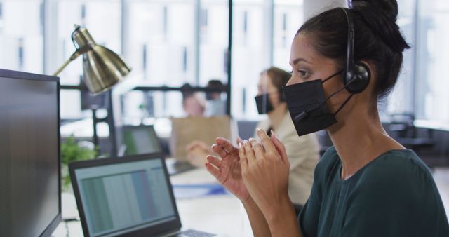 Customer service agent wearing face mask and headset working at office desk while using computer. Suitable for illustrating concepts related to customer support, COVID-19 safety measures, professional office environments, remote assistance, and work during pandemic conditions.