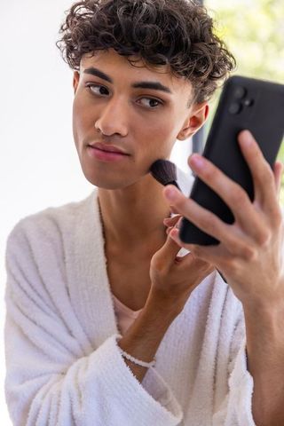 Young man applying powder makeup using phone at home
