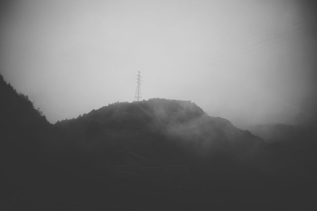 Fog enveloping a mountain hill with an electricity tower in the distance, creating a misty and mysterious atmosphere. Ideal for backgrounds, nature-themed projects, environmental awareness materials, and minimalist design implementations.