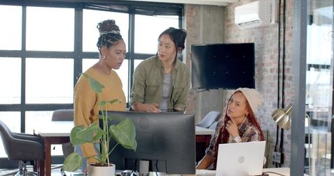 Three women collaborating at dual monitors in modern urban loft office