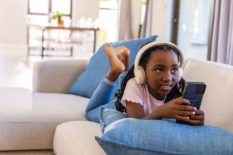 School-age african american girl relaxing on sofa with headphones and handheld device