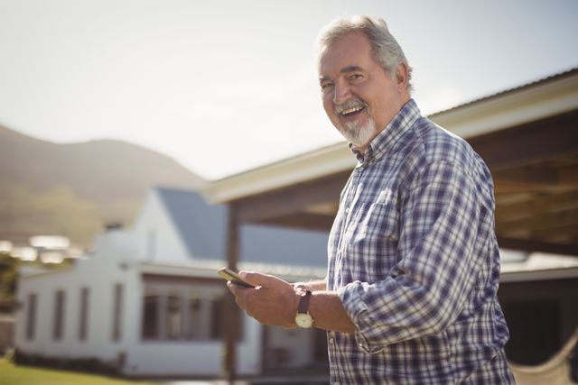 Portrait of smiling senior man with his mobile phone standing outside his house
