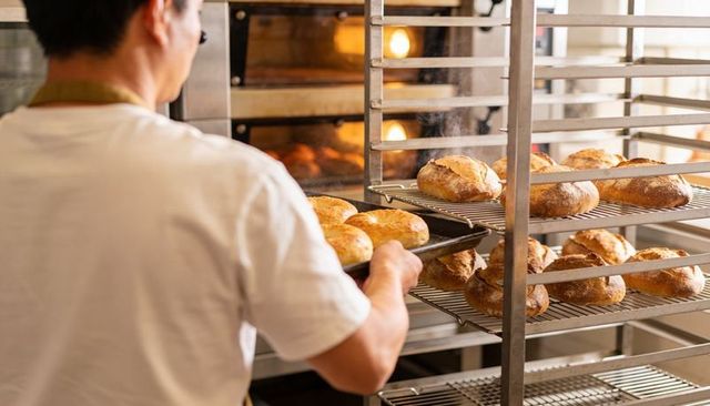 Baker Placing Tray of Fresh Artisan Loaves onto Cooling Rack Next to Commercial Oven