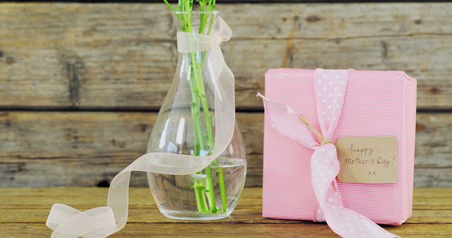 A pink-striped gift box tied with a ribbon and a tag saying Happy Mother's Day is placed next to a clear vase with green stems, set against a rustic wooden backdrop. The scene evokes a warm, celebratory atmosphere for Mother's Day, highlighting thoughtful gifting and the beauty of simplicity.
