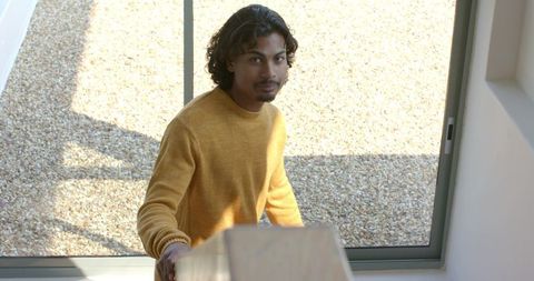 Asian man standing on sunlit modern stairwell wearing yellow sweater touching banister