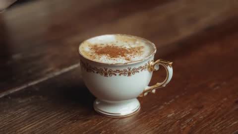 Pouring dark coffee stream creating cinnamon-dusted foam in ornate porcelain teacup on wooden table