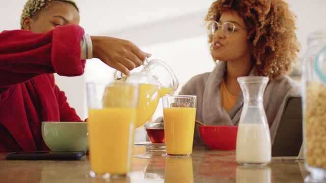 Two diverse young women sitting at a kitchen table enjoying a breakfast of cereal and orange juice. One woman is pouring juice while the other is engaging in conversation, both dressed in casual attire, emphasizing a comfortable and relatable domestic scene. Suitable for use in content related to friendship, home life, morning routines, and quality time indoors.