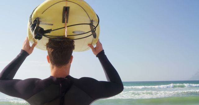 A young Caucasian man in a wetsuit holds a surfboard over his head, ready to tackle the waves, with copy space. His anticipation for the surf session is palpable as he gazes towards the sea.