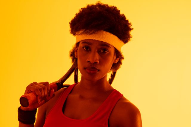 African american female tennis player with afro hair holding racket against neon yellow background. Copy space, portrait, confident, sport and competition concept.
