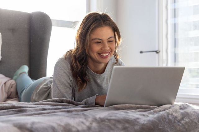 Smiling woman lounging on bed using laptop for remote work and online study in bright apartment