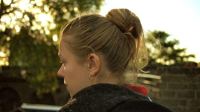 A young woman with blonde hair tied in a ponytail relaxing outside during sunset. The image captures her from the back, presenting a thoughtful and peaceful atmosphere. It can be used for themes of relaxation, tranquility, and the beauty of nature during sunset.