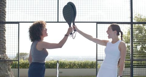 Diverse women tapping paddles on sunlit outdoor padel court celebrating teamwork