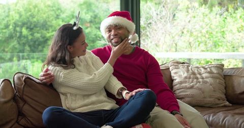 Diverse couple sharing cozy christmas moment on sofa wearing santa hat and antlers